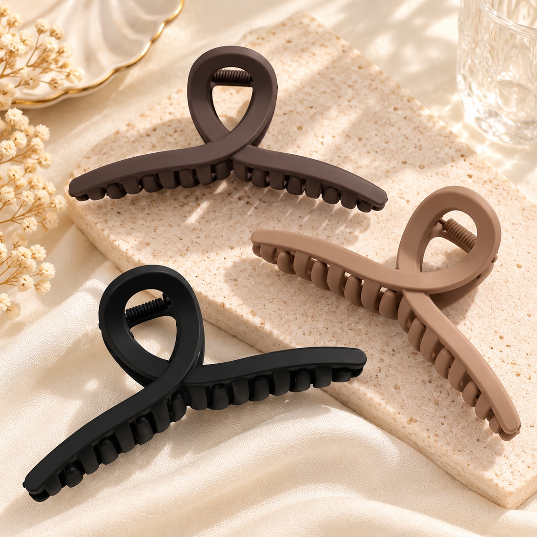 Three minimalist loop-shaped matte hair claw clips in black, taupe and brown arranged on a beige surface